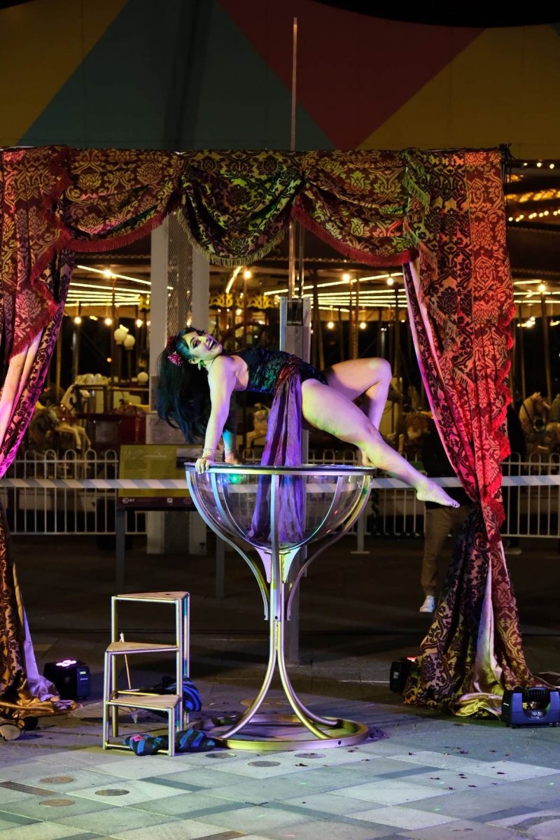 Woman dancing on top of a giant martini glass with curtains and a carousel in the background.