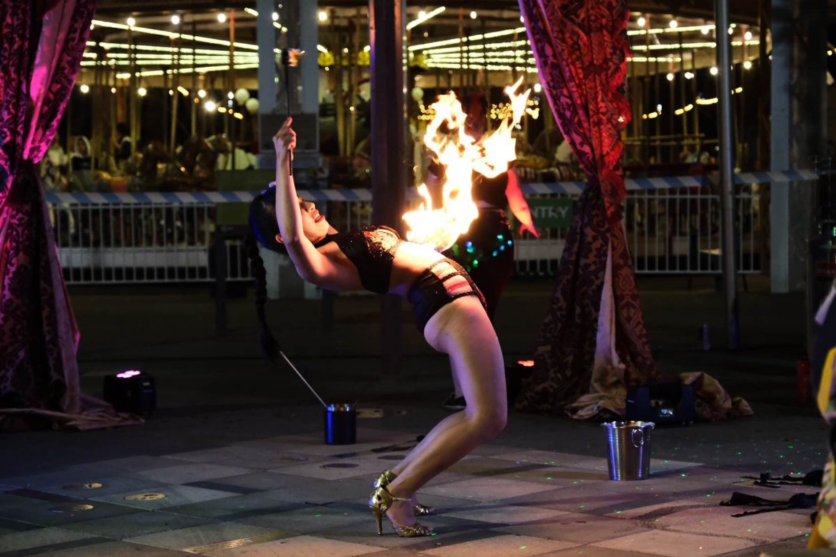 Woman arching her back with a flame coming off her belly with a set of open curtains revealing a carousel in the background.