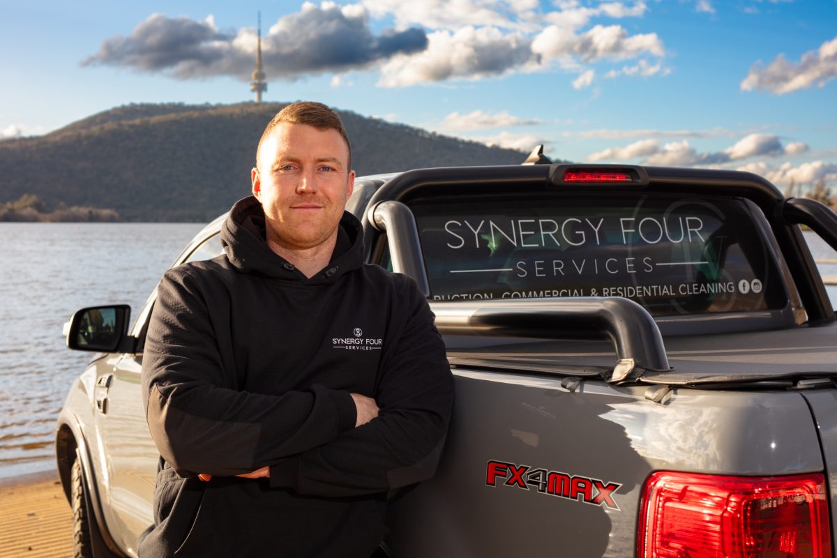 Man standing in front of a car at a lake, with Telstra Tower in background.