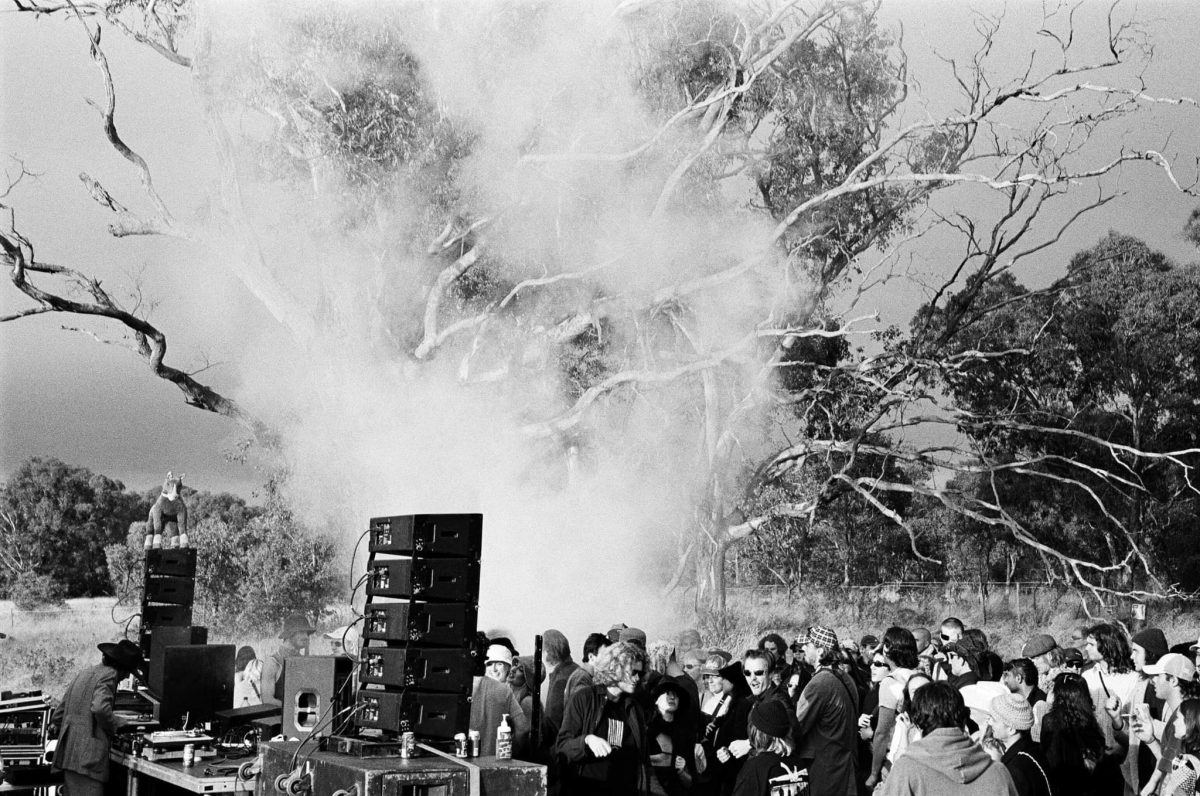 A black and white image of a DJ set in the bush with gumtrees in the background and a group of people dancing in front with fake smoke swirling up in the middle.