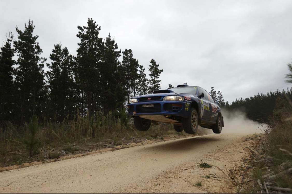 A subaru rally car in mid air while racing on a dirt track.