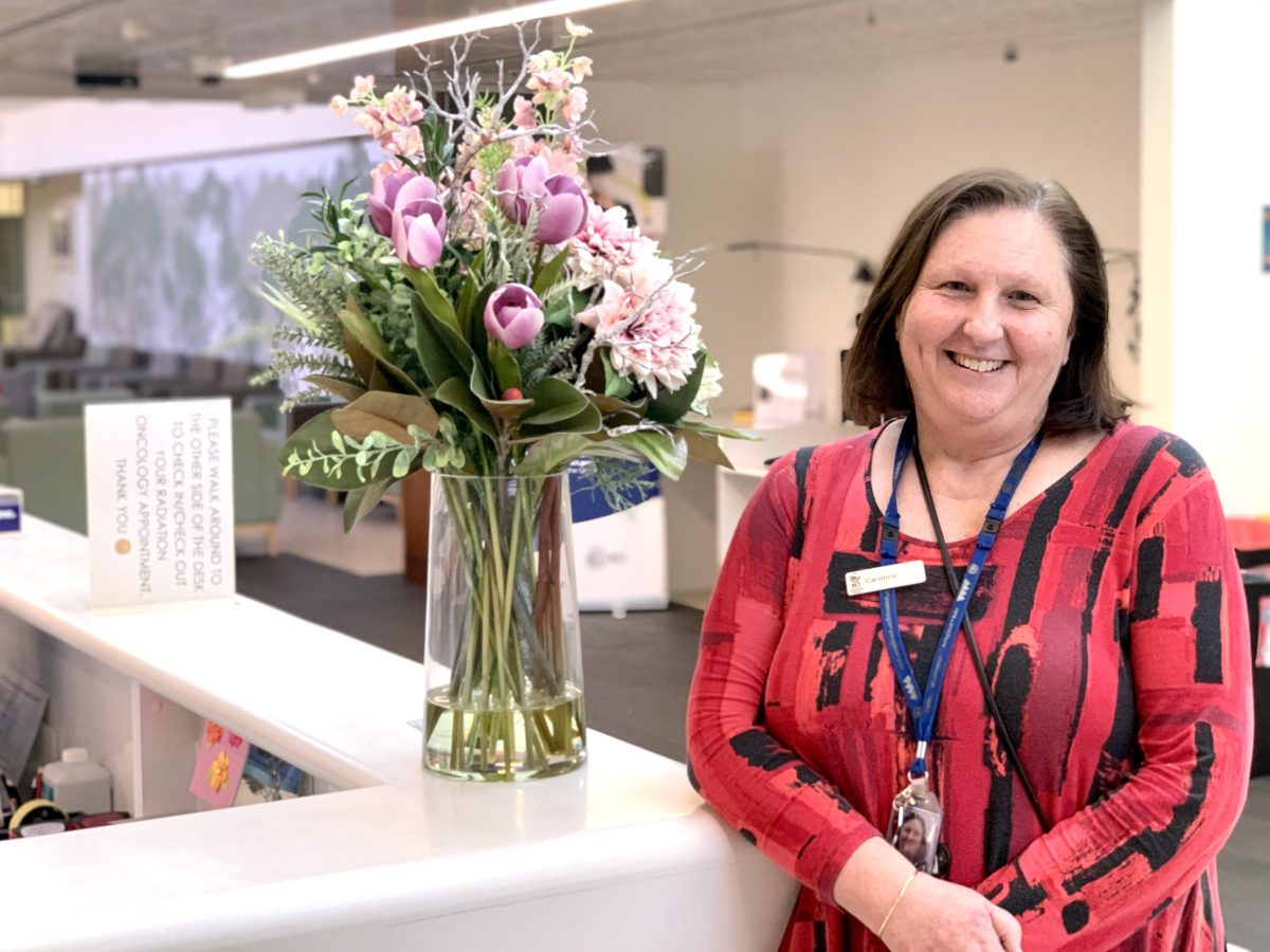 Caroline McIntyre smiles and leans on a hospital reception counter, next to a bunch of flowers