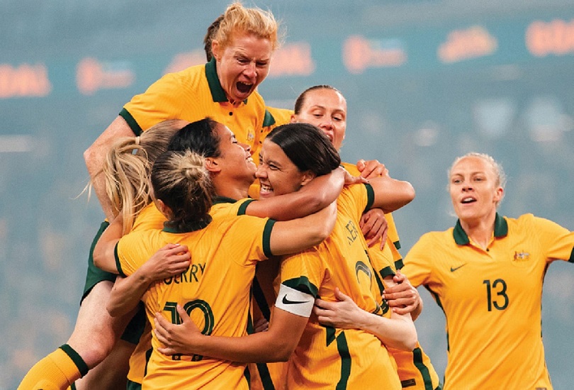 The Matildas celebrating in a huddle. One woman is jumping into the huddle.
