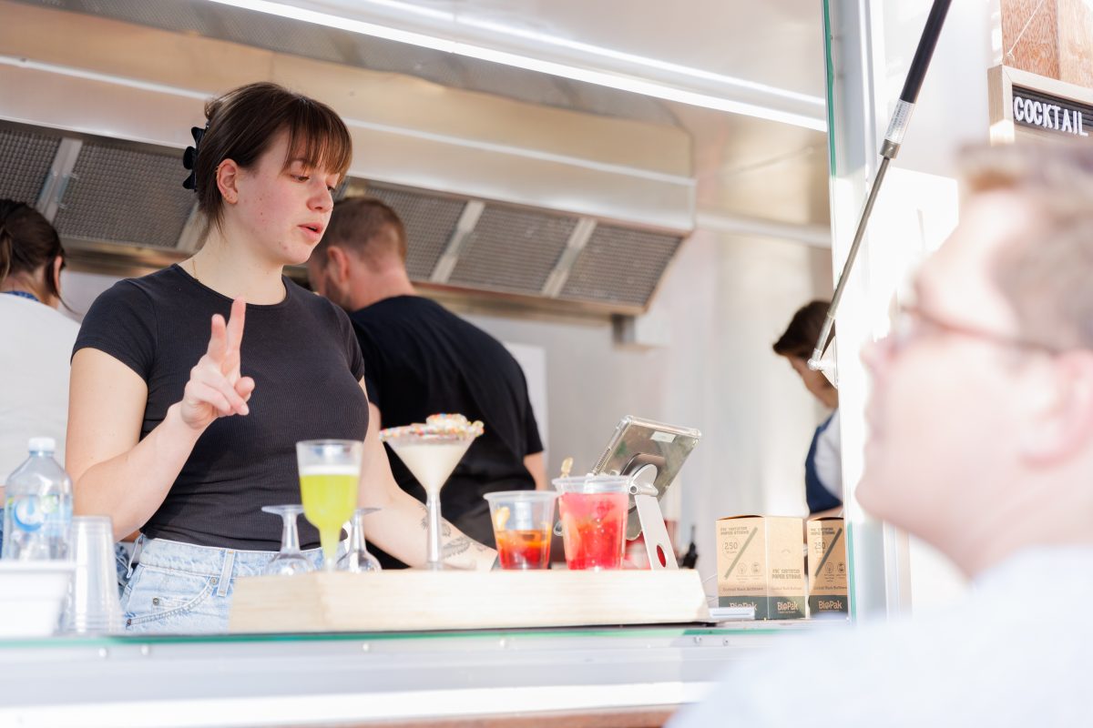 Inside a truck, a bartender takes orders with a line of cocktails in front of her, and a customer in the foreground.