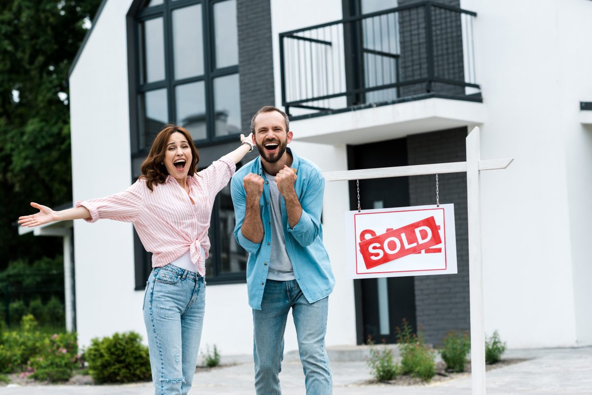 cheerful man and woman celebrating near house and board with sold letters
