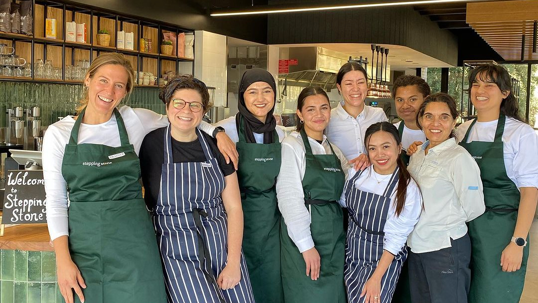 Group of women in aprons smiling in front of cafe counter.