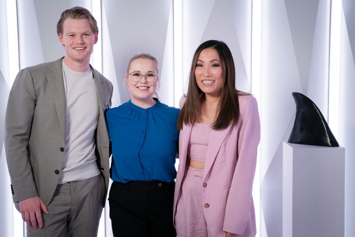 Man in grey suit, woman with blue top and woman in pink suit stand in front of a lit background with a shark fin trophy next to them.