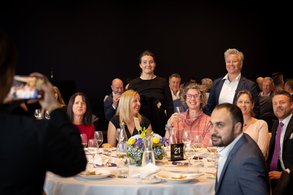People seated around a table set with white table cloth, plates and glasses - with two people standing behind 2022 Philanthropy Award Winner Jo Farrell.