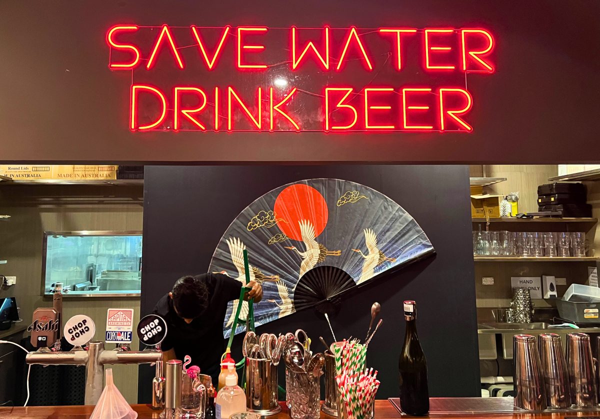 Neon sign "save water drink beer" and large decorative fan behind a bar