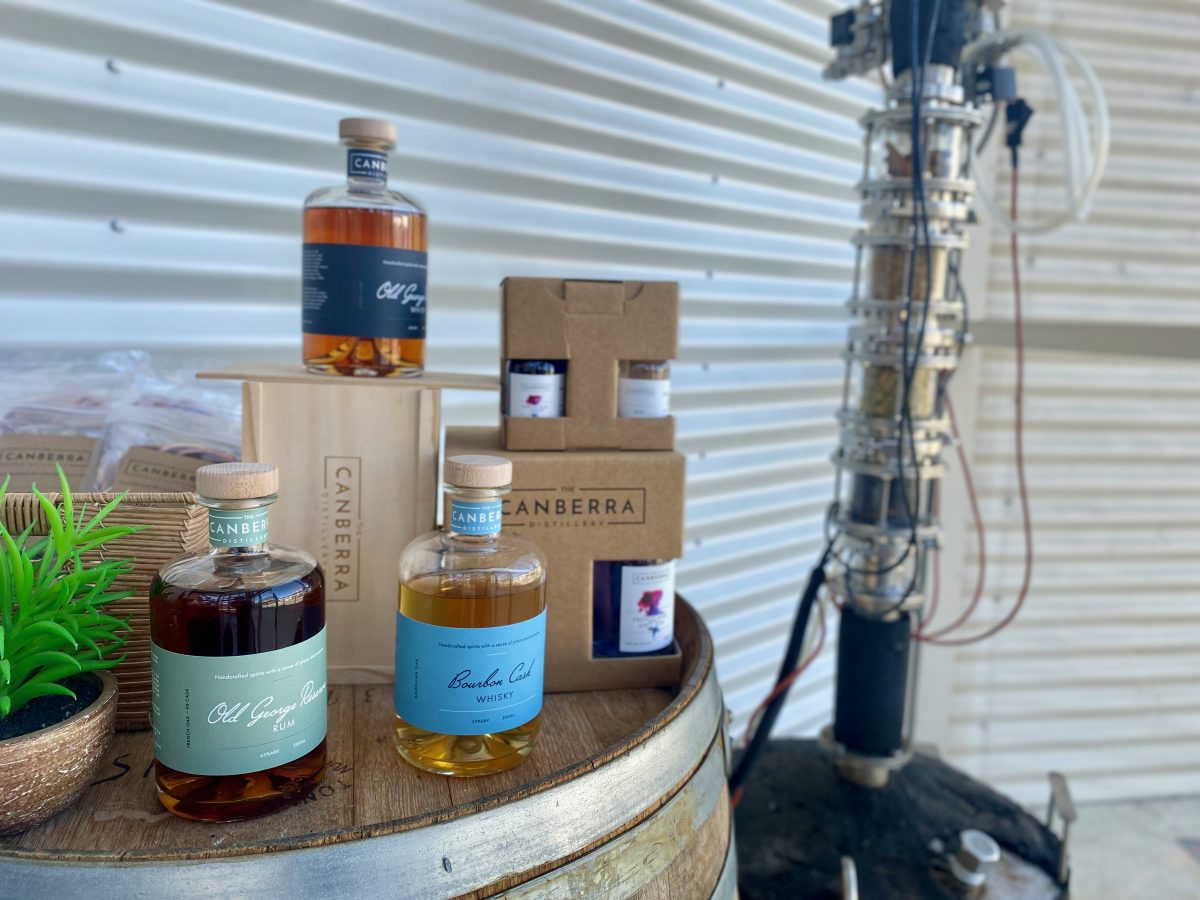 Bottles of whisky and rum on a barrel with distilling equipment in the background.