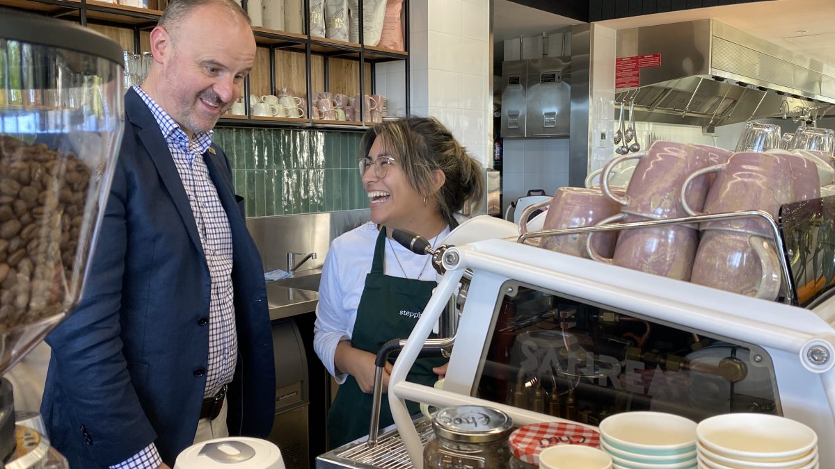 Man in suit stands next to woman in apron behind a coffee machine.