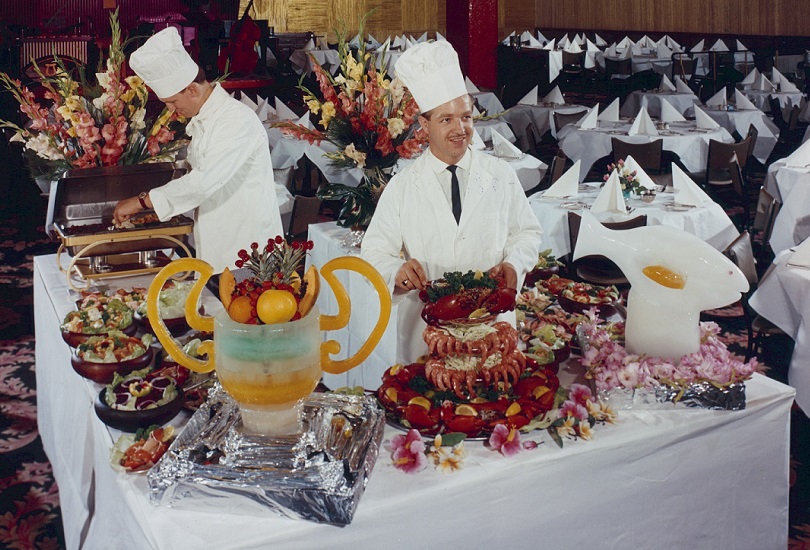 Two large white tables filled with food and drink, two chefs behind the tables. 