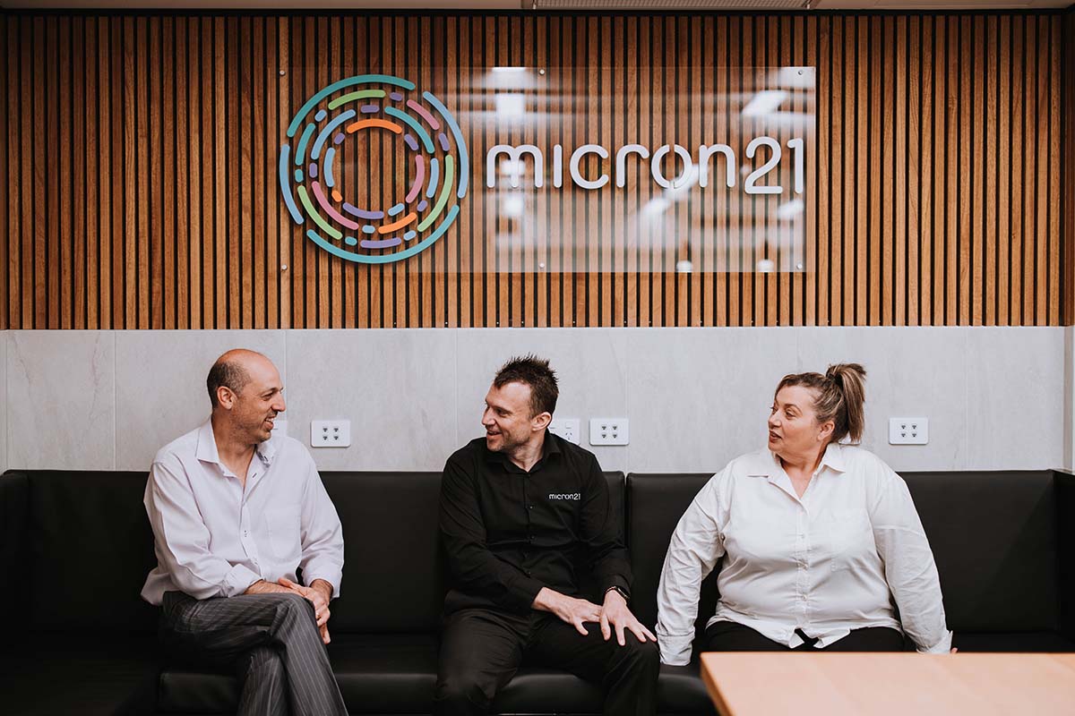 The team at Micron21 Sheldon Dyer, James Braunegg and Michelle Anderson sitting on a lounge under a Micron21 sign. 