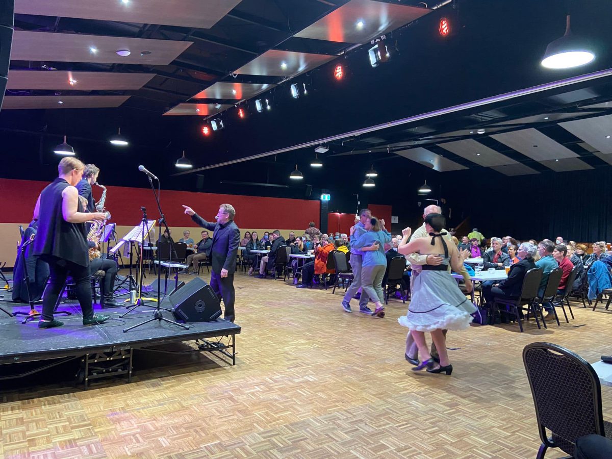 Band (left) playing to a crowded room of seated patrons (right) who are watching a two couples dance in between them. 