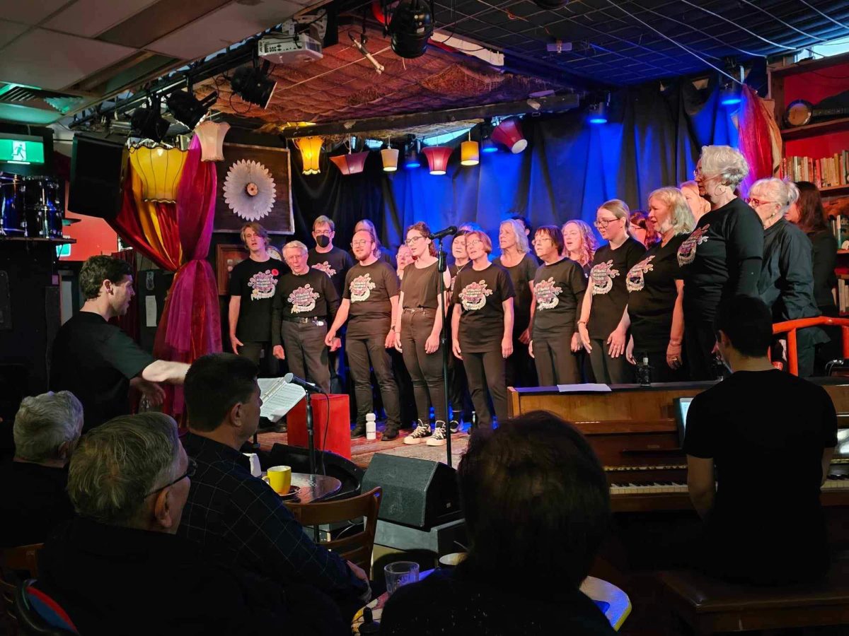 The choir performing at Smith's Alternative Cafe with piano playing beside them and a crowd sitting in the foreground.