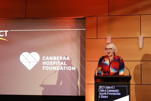 Kate Palmer standing at lectern in room with red lighting and Canberra Hospital Foundation logo on a screen.