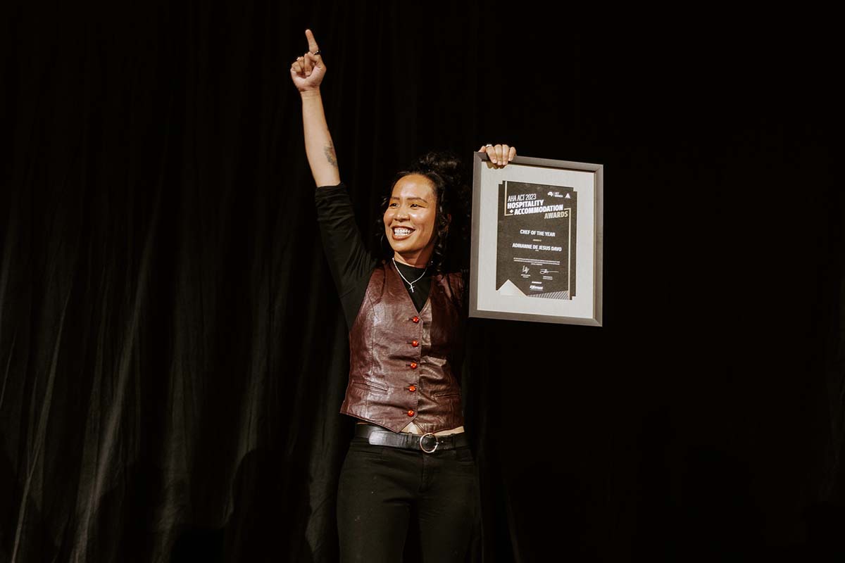 Adrianne wears a brown vest and points up with one hand while holding her framed certificate reading "Chef of the year" in the other