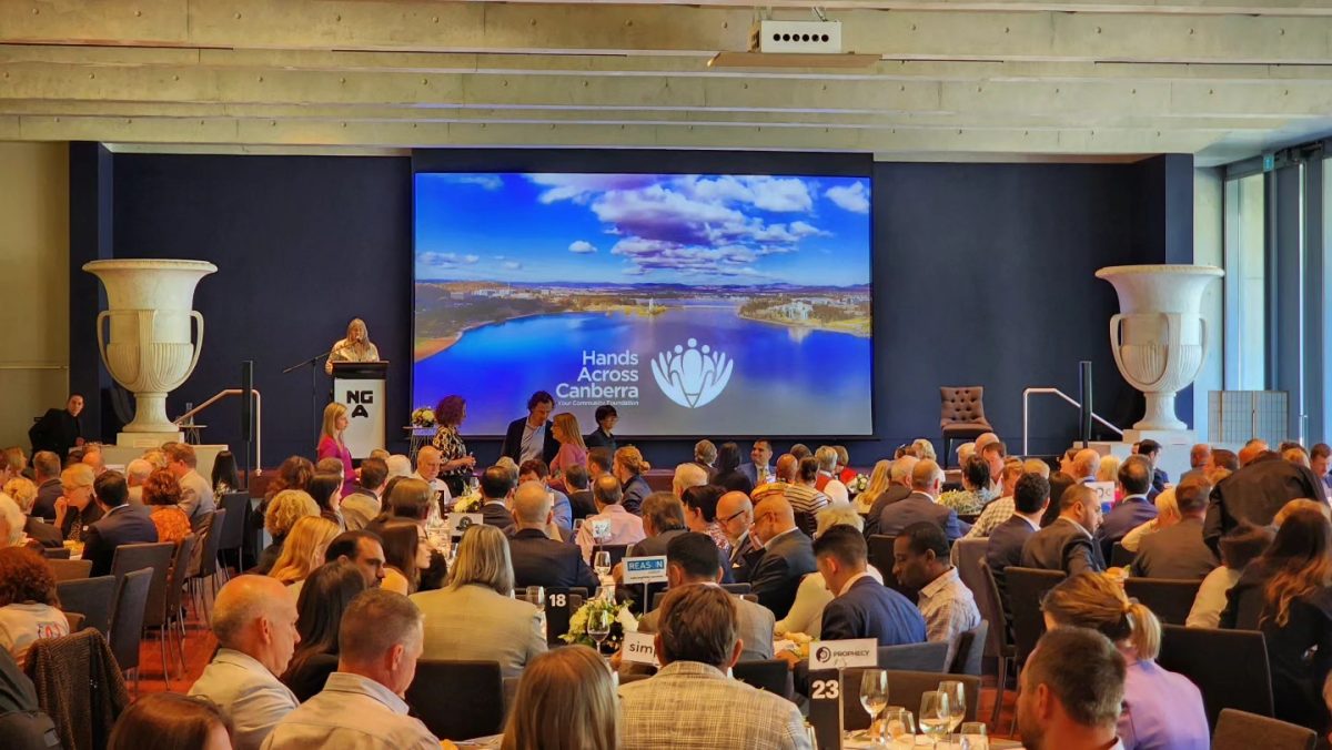 Room filled with people sitting at tables with a large screen showing the Hands Across Canberra logo and photo of Canberra