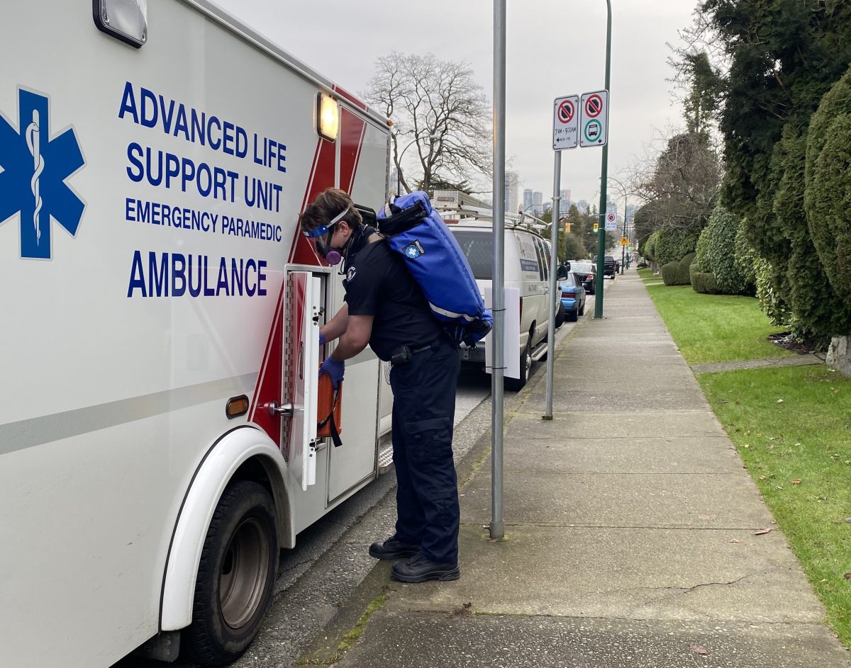 Richard in paramedic uniform with a bulky blue backpack, gas mask, and transparent visor on, grabbing something from the side of an ambulance parked on the street in Vancouver.