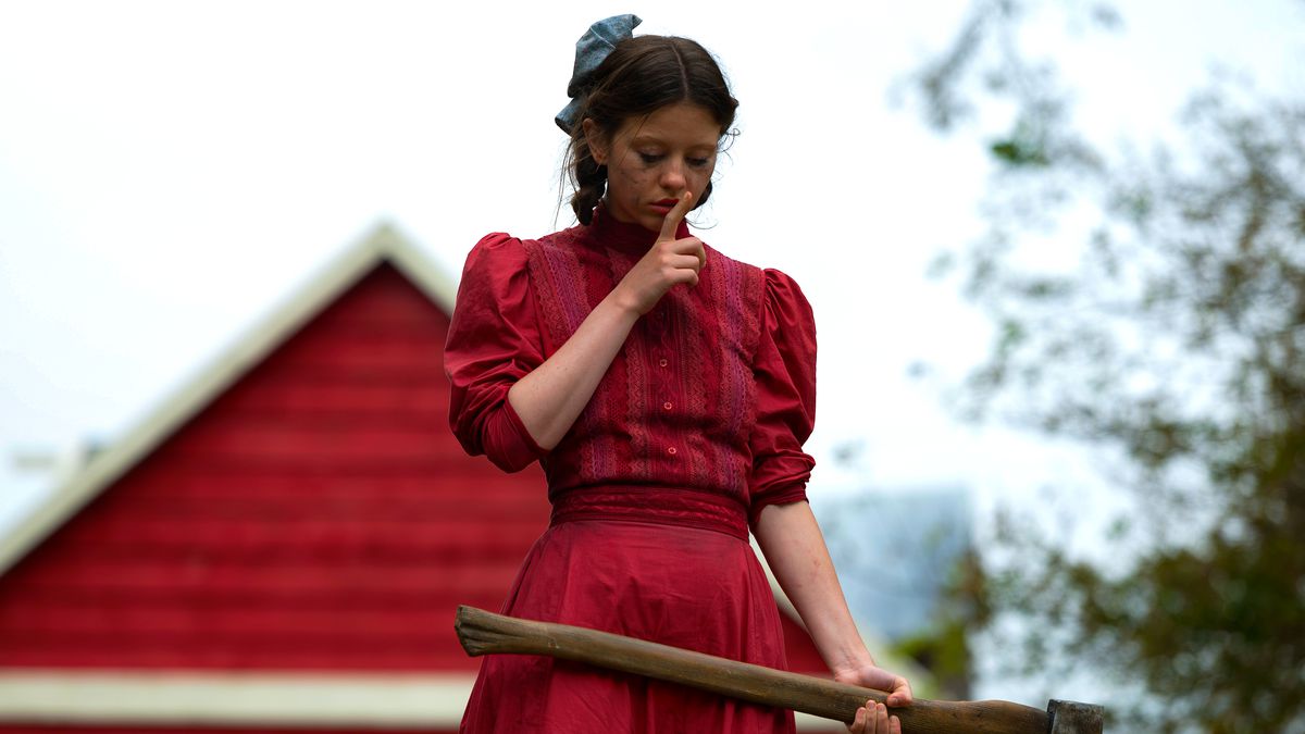 Mia Goth in a red dress holding an axe with a red barn behind her