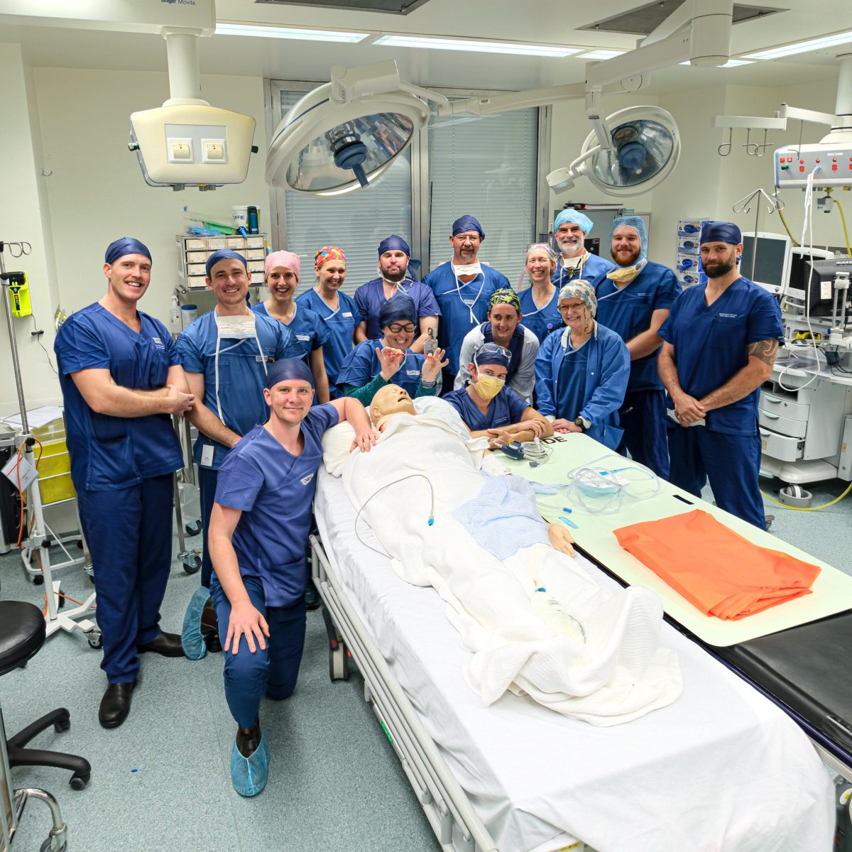 A group of students in medical gear standing around a plastic body on the bed inside an operating room.