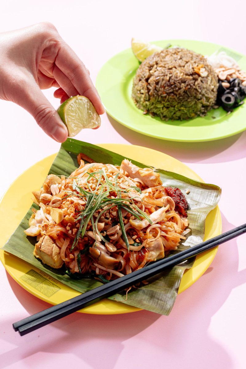 A white hand squeezes a wedge of lime onto a noodle dish, in the background a plate with a mound of rice and cut black olives