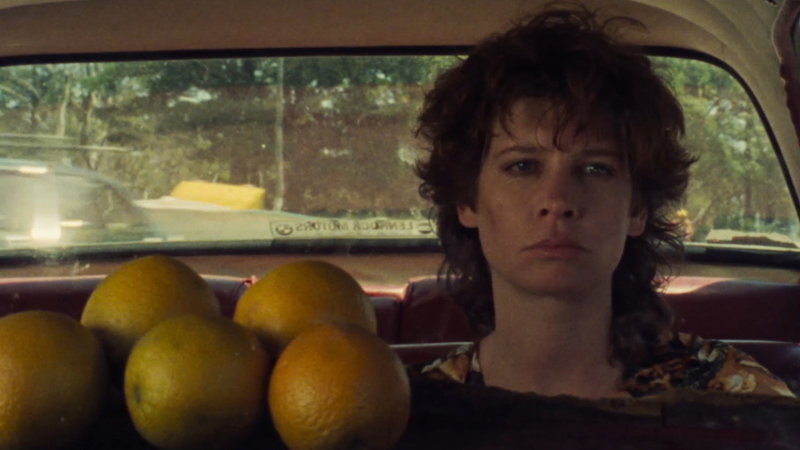 A still from the movie Peel of a woman sitting in the car with a few oranges on the dashboard and a leafy background seen out of the car's rear.