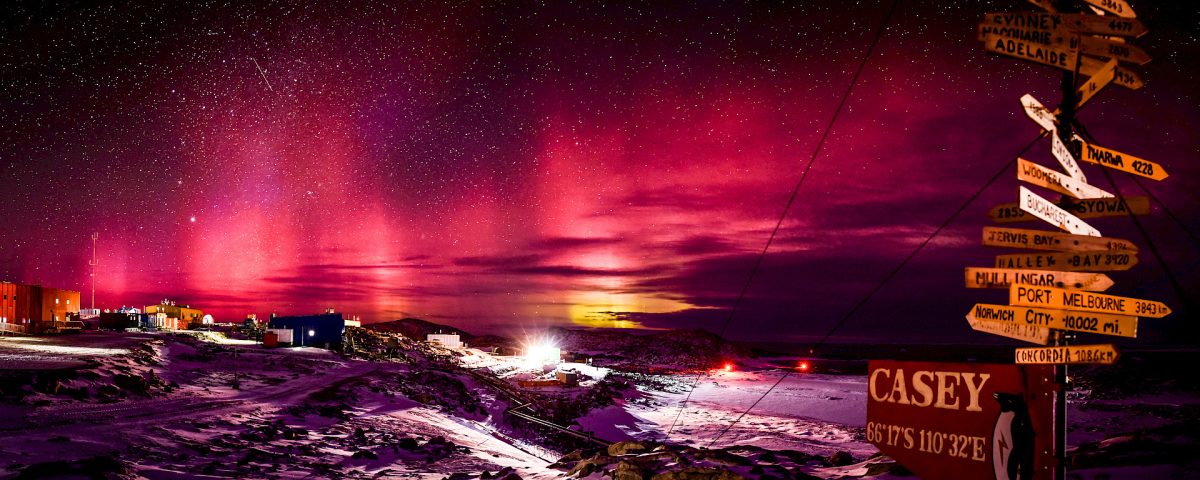 An aurora at night time over Casey Station in Antarctica with a pole holding stacks of signs pointing to places all over the world.