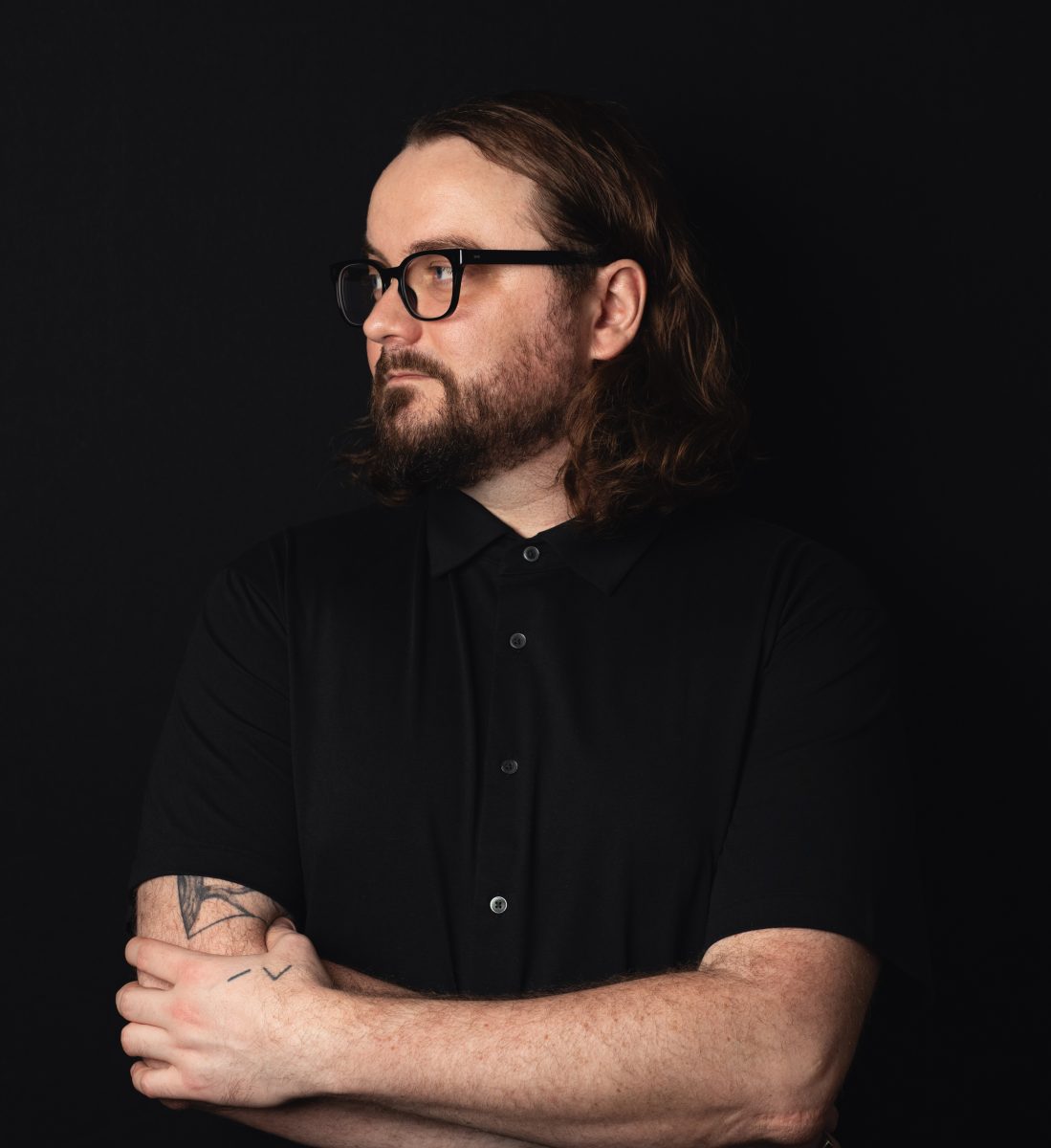 A white man with long dark hair and beard wears black glasses, black shirt against black background.