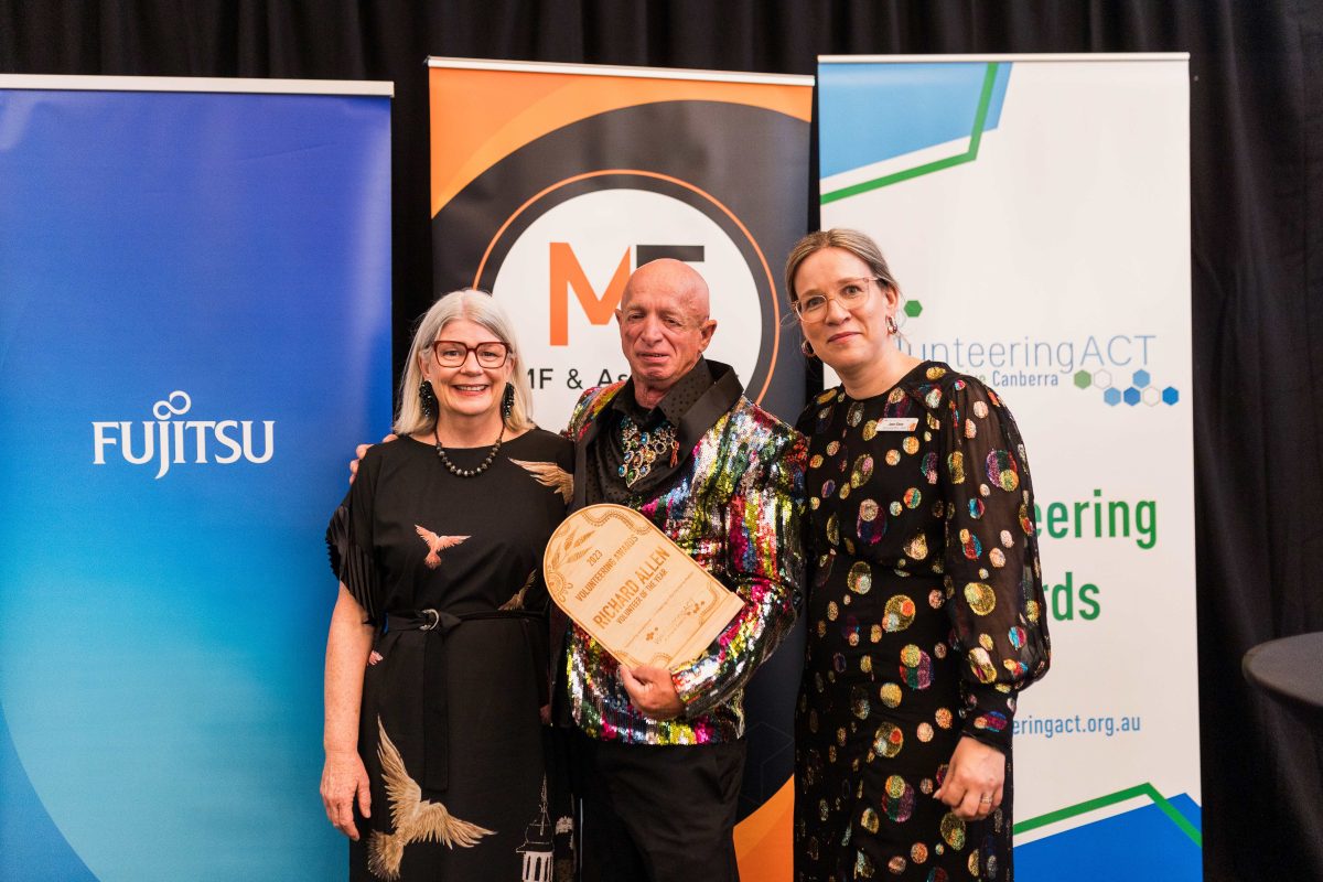 Two women next to man in shiny jacket holding award.