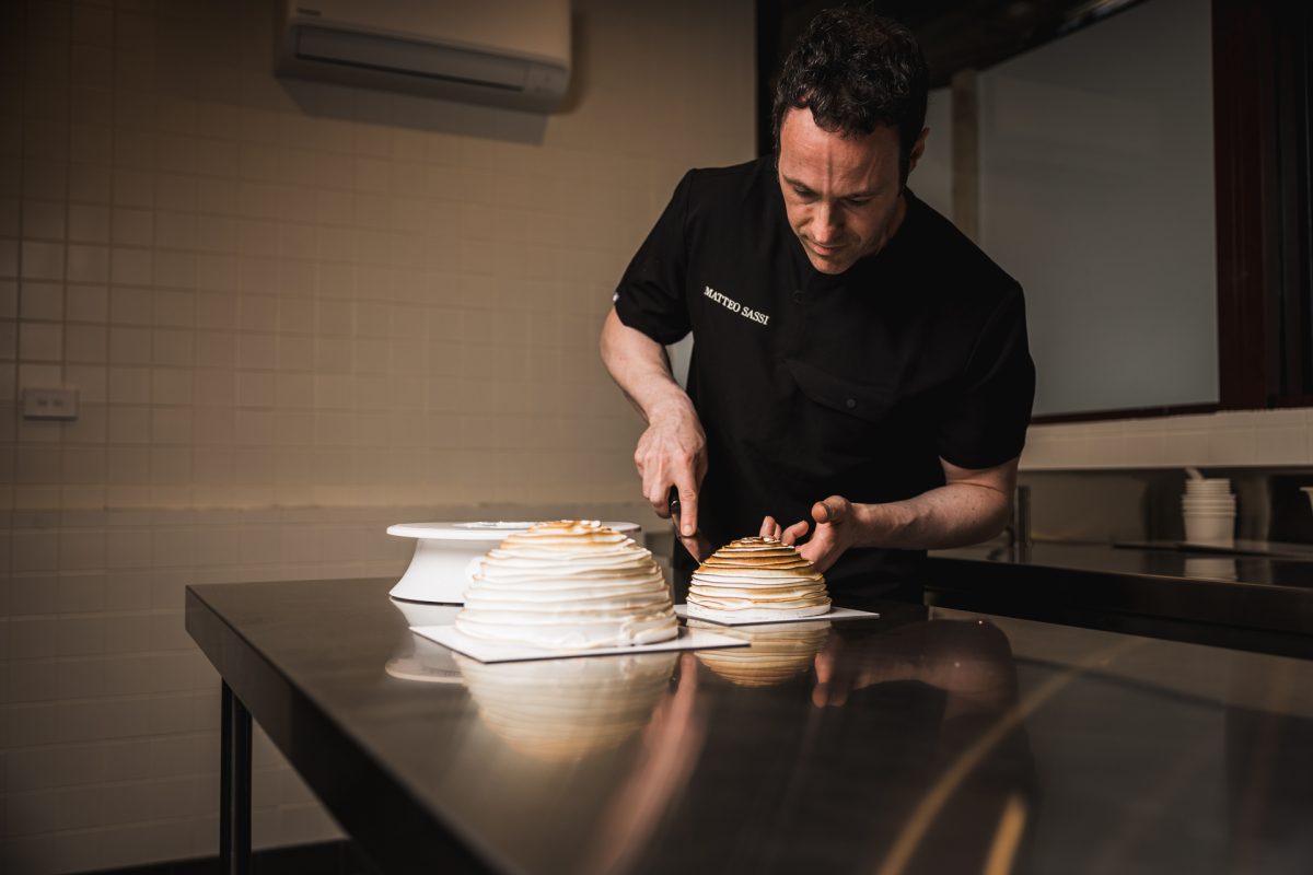 Man in black shirt with white text reading ''Matteo Sassi'' ices a cake