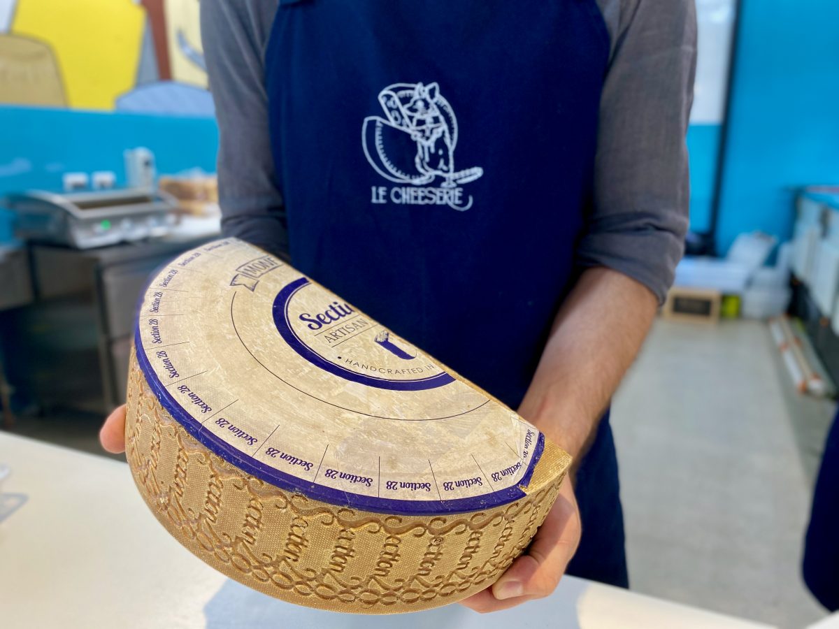 A large half wheel of cheese with Section 28 stamped on rind is held, Le Cheeserie logo is on the apron in the background.