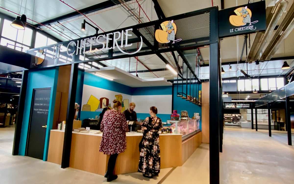 Exterior of Le Cheeserie store in the market. Two customers are at the counter.