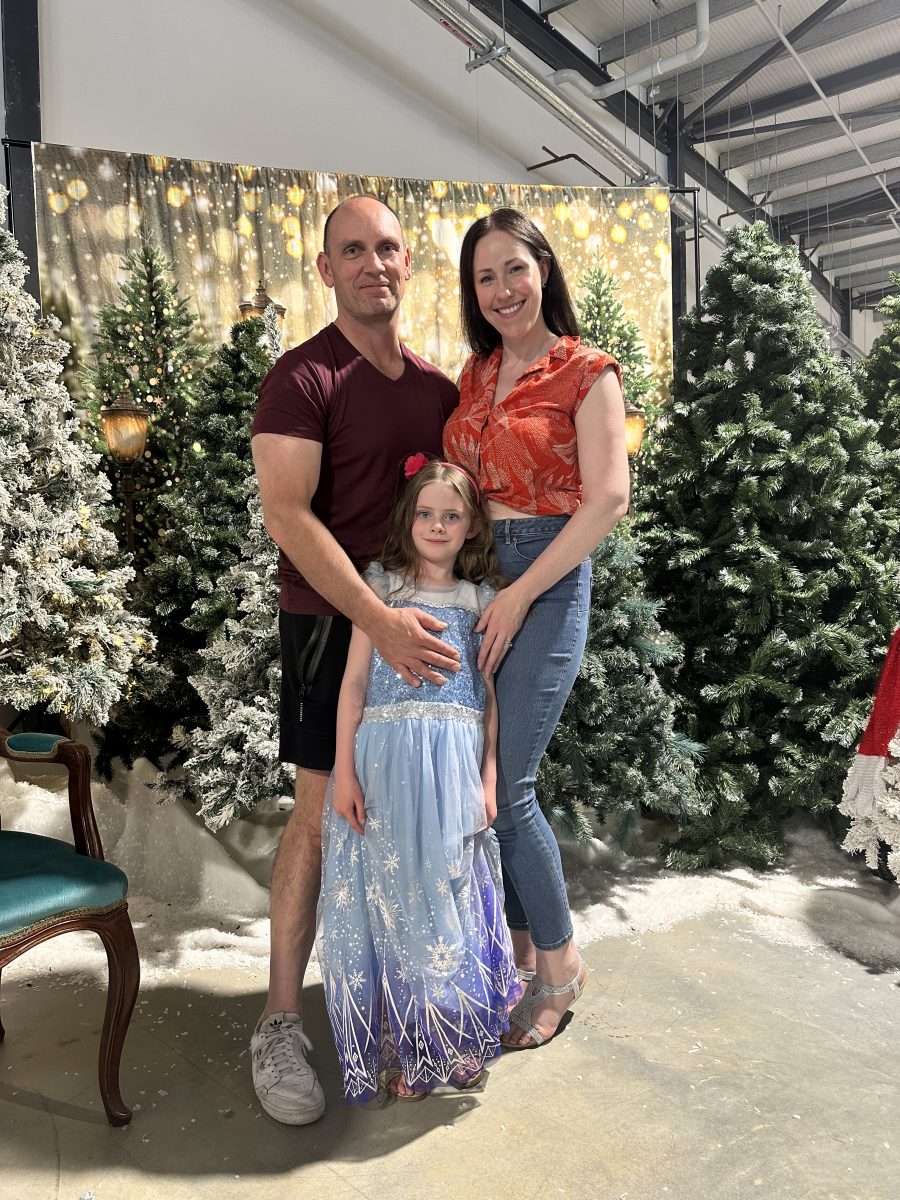 Man, woman and little girl in front of Christmas trees. 