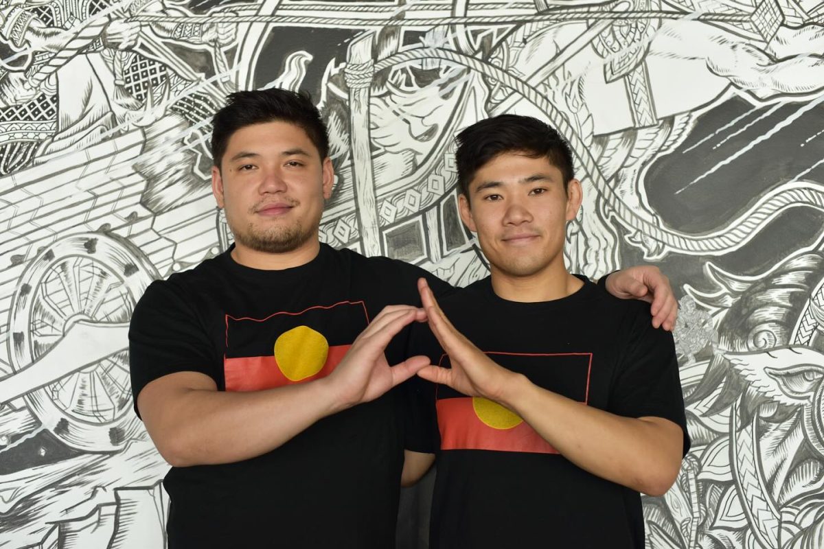 Two men stand if front of a black and white graffiti wall. They wear Aboriginal flags on their shirts and are making the shape of Australia with their hands