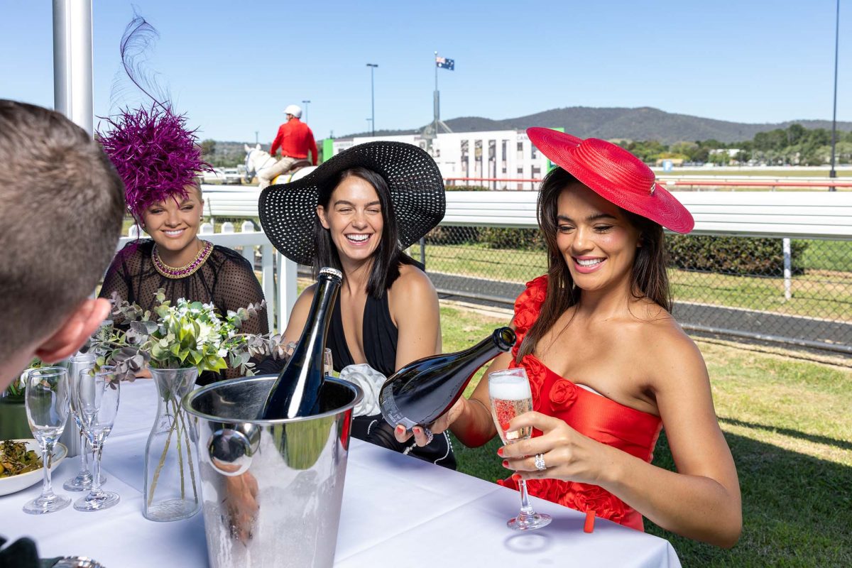 three women sitting at a table at the races drinking champagne