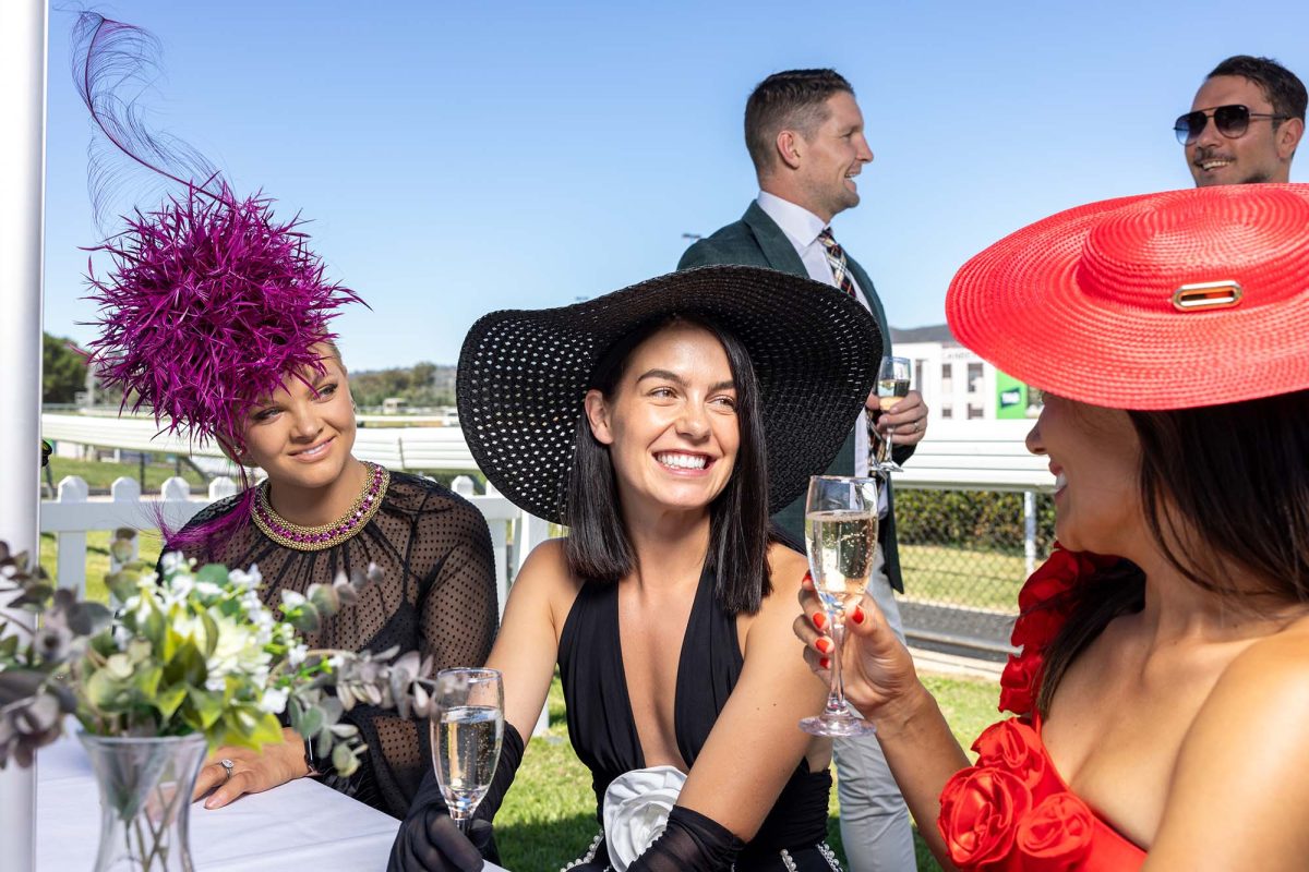 three women wearing fascinators sipping champagne trackside at horse races