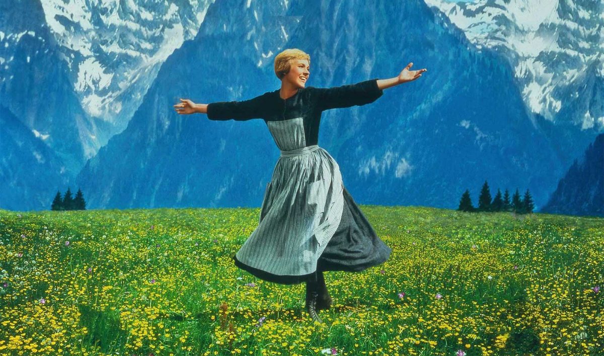 Still from the film The Sound of Music showing a woman spinning in a green meadow. There are mountains in the background.
