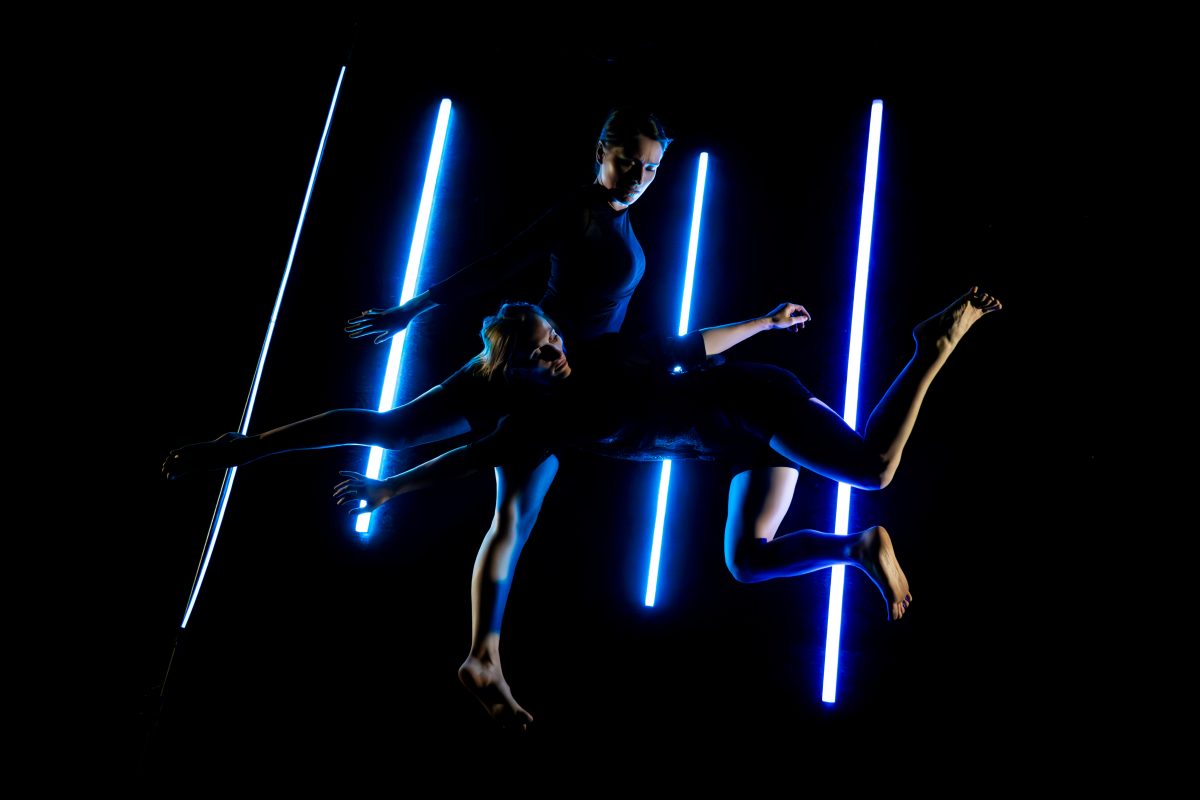 Two dancers in black pose in front of neon light display.