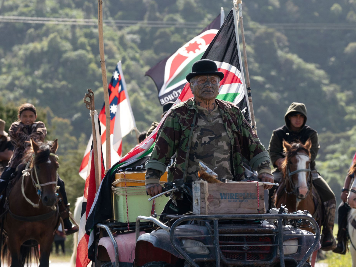 Still from Muru showing a man on a quad bike leading a group of people on horseback with flags