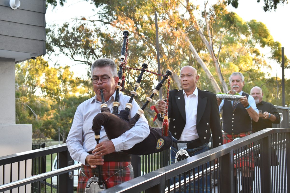 Four men march over a bridge in Scottish kilts, the leader playing bagpipes