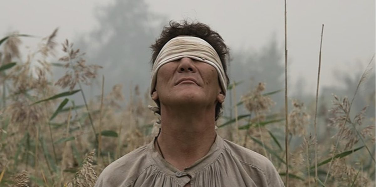 Still from Don't Bury Me Without Ivan showing a blindfolded man looking up in a field