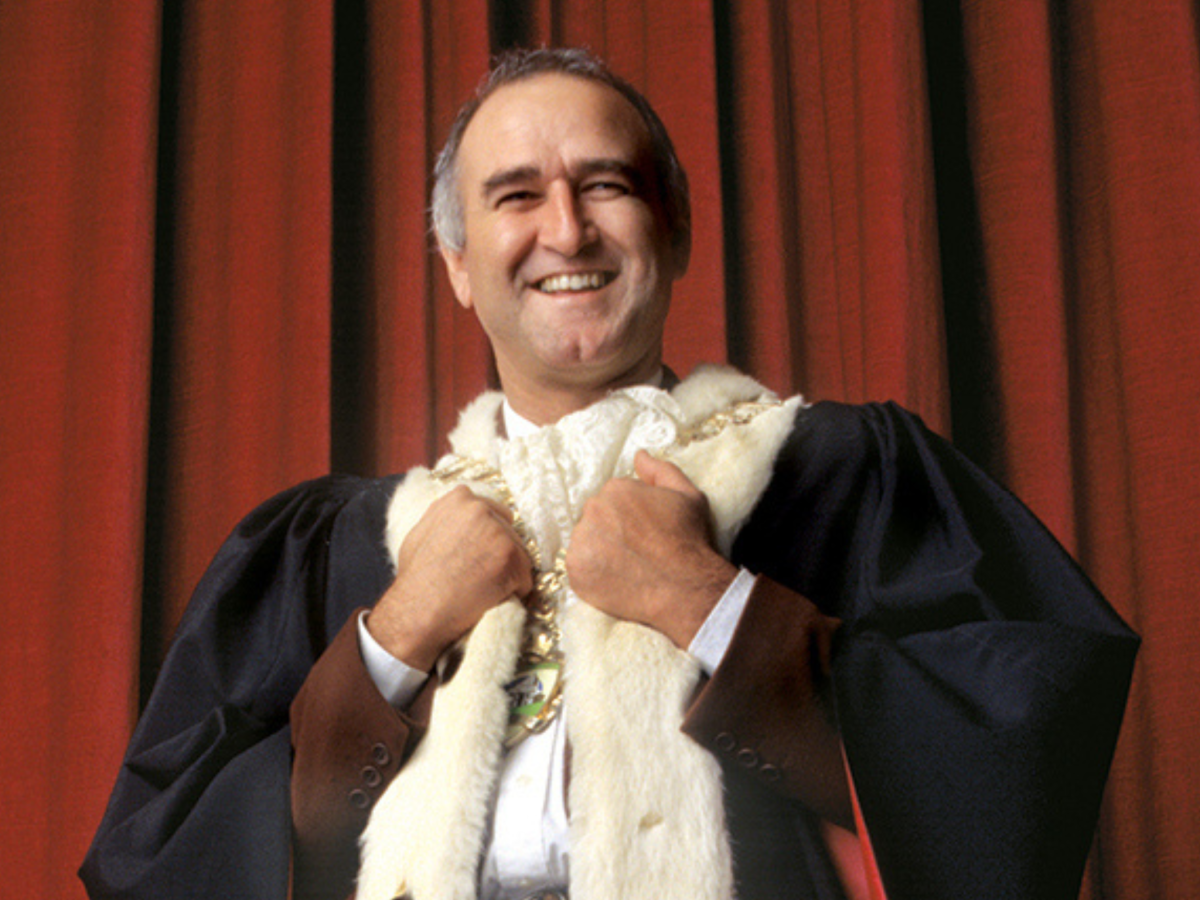 Promotional image for Rats in the Ranks showing a man wearing a fur coat, standing in front of a red curtain