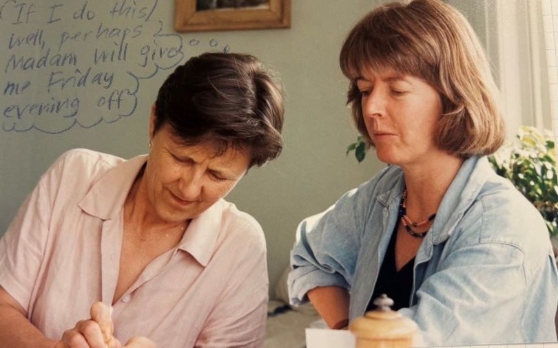 A photograph of two women concentrating on the work of the woman on the left. The woman on the left has a drawn thinking bubble above that says, 'If I do this well, perhaps Madam will give me Friday evening off.'