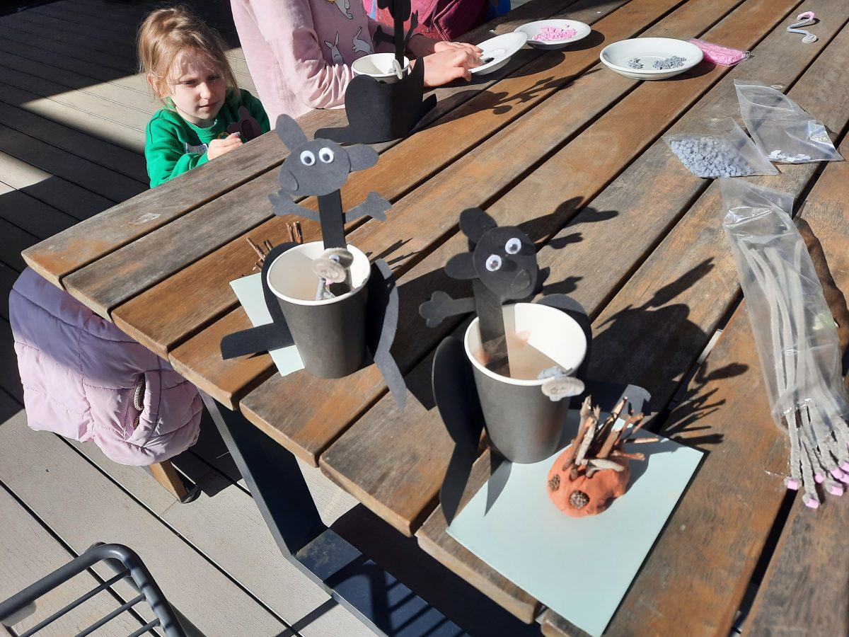 Two black wallaroos made from paper cups and carboard are sitting central on a wooden table with an echidna made of clay and stick to the right of them.