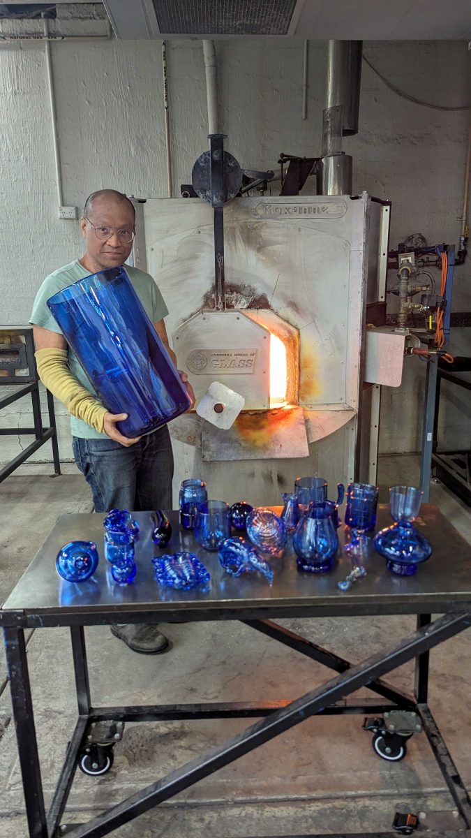 Man near kiln with table of blue glass objects
