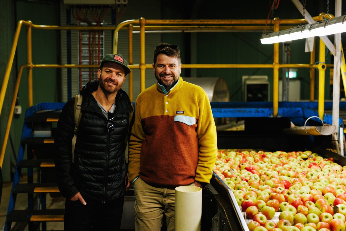 Two men stand next to a large vat of apples