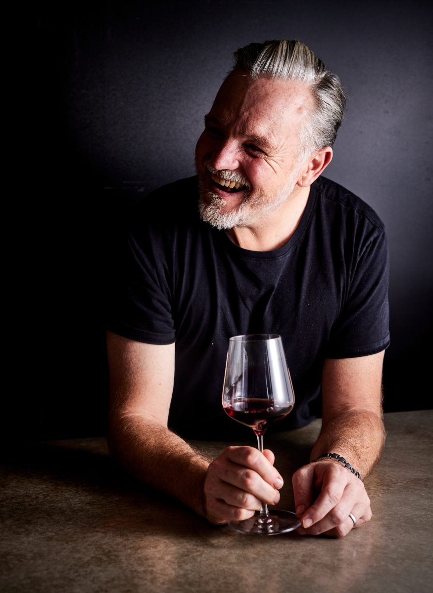 Man with grey hair and beard laughs while holding wine glass.