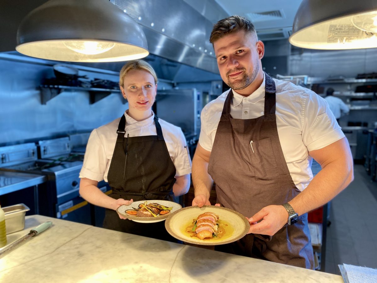 Two chefs hold plates at the kitchen pass. 