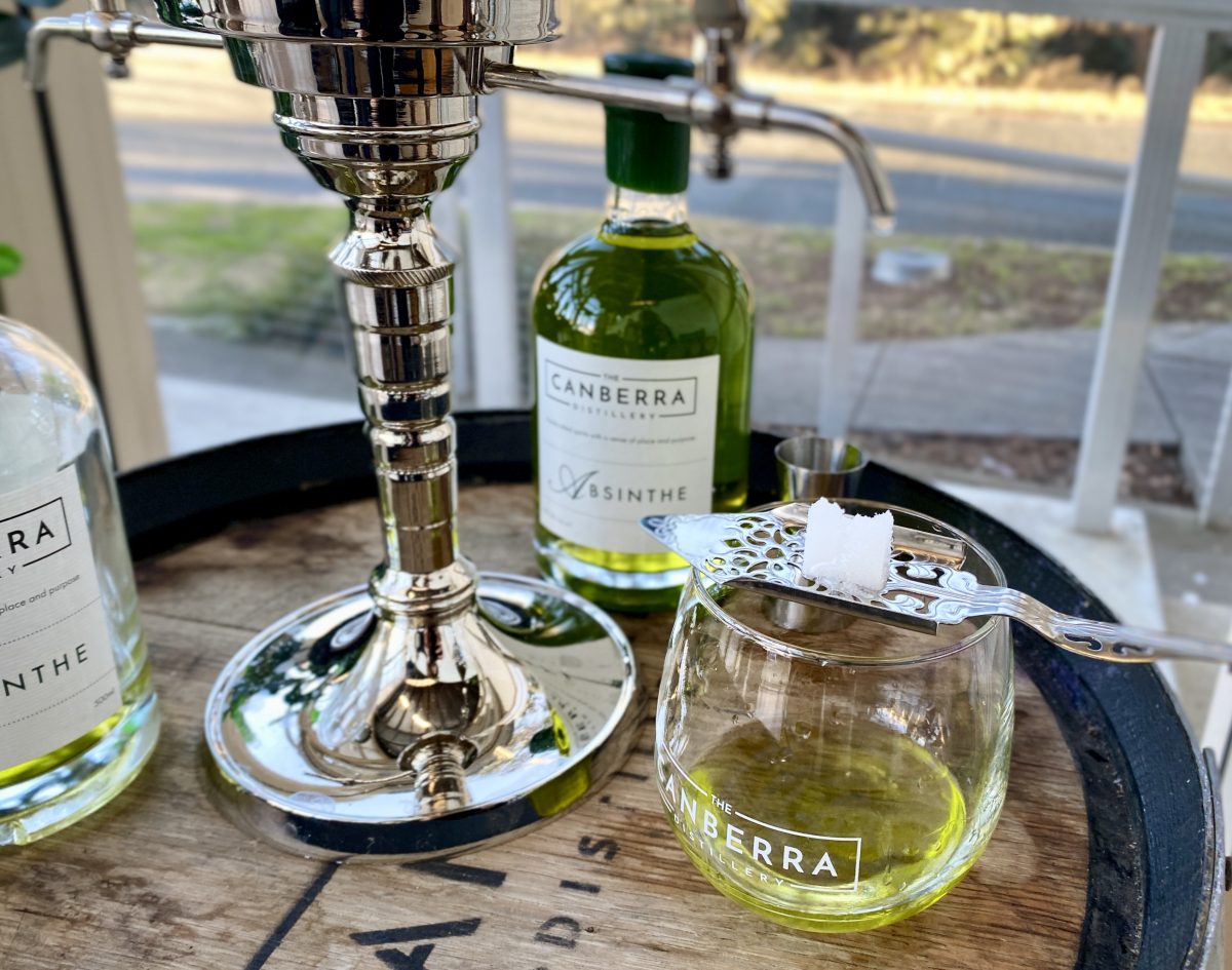 An absinthe fountain with sugar cube balanced on flat, perforated silver spoon over a Canberra Distillery branded glass.